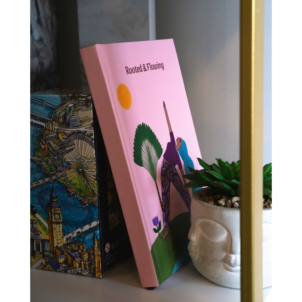 Load image into Gallery viewer, A pink hardcover book titled 'Rooted & Floating' with a colorful illustration on the cover, leaning against a box with a London-themed design next to a small potted succulent.