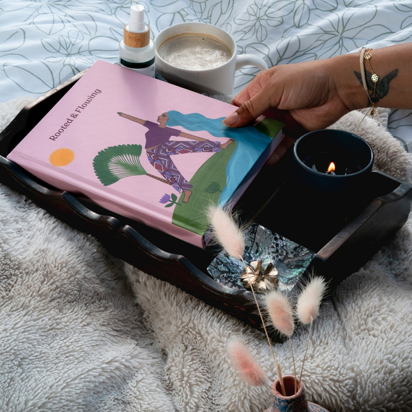 Load image into Gallery viewer, A hand holds a pink book titled 'Boundless & Blooming' on a dark wooden tray, styled with a mug of coffee, a lit candle, and dried bunny tail grass on a fluffy gray blanket.