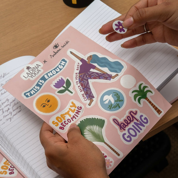 Load image into Gallery viewer, A person's hands peel and display colorful stickers from a sheet titled 'This Is Enough' by Laadna Yoga x Melodicalyze. The sheet features various designs including a yoga pose, a purple flower, a smiling sun, and motivational quotes on a pink background, shown over an open lined notebook on a wooden desk.