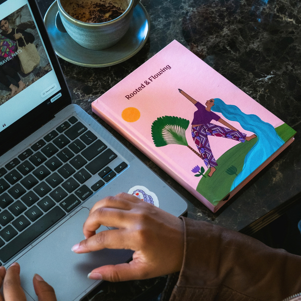 Load image into Gallery viewer, A pink hardcover book titled 'Rooted &amp; Flowing' with a colorful illustration of a woman dancing, placed on a dark marble desk next to an open laptop and a cup of tea. A person's hands are visible typing on the laptop.
