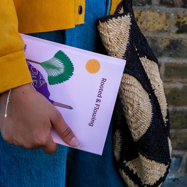 Load image into Gallery viewer, A person in a yellow jacket and blue jeans holds a pink notebook titled 'Rooted &amp; Flourishing' next to a woven black and beige shoulder bag, with a brick wall in the background.
