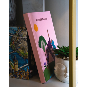 A pink hardcover book titled 'Rooted & Floating' with a colorful illustration on the cover, leaning against a box with a London-themed design next to a small potted succulent.