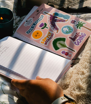 A pink sticker sheet with colorful motivational and tropical stickers is placed over an open lined notebook on a soft cream rug. A hand holds the notebook, and a lit candle and patterned blanket are visible in the background.