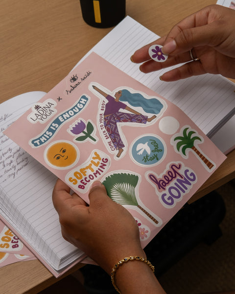 Load image into Gallery viewer, A person's hands holding a pink sheet of die-cut stickers with various designs, including a cartoon figure, palm trees, and motivational phrases, over an open notebook on a wooden desk.
