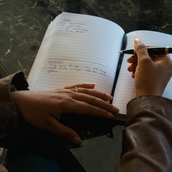 Load image into Gallery viewer, A close-up of a person writing in an open lined notebook with a black and gold pen, resting on a dark marble surface.

