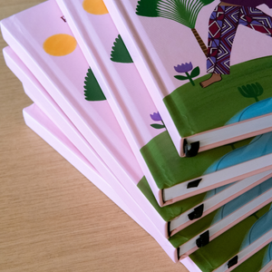 A stack of hardcover notebooks with pastel pink covers and colorful illustrated designs, photographed on a light wood surface.