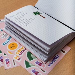 An open lined notebook with handwritten notes and a small palm tree illustration sits on a wooden surface, layered over pink sticker sheets with tropical and beach-themed designs.
