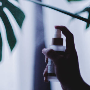 Woman's hand holding a yoga mat spray made with Ylang Ylang essential oil to clean a yoga mat. Background are green plants